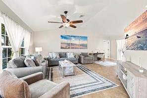 A spacious living room with a ceiling fan, a large painting, and a view of the outdoors. - The Dock House I Gulf Access I Southern Exposure Pool/Spa (Cape Coral)
