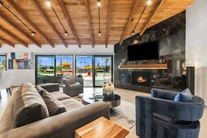 A modern living room with a fireplace, a large TV, and a view of a pool and palm trees. - Stunning South Palm Desert Home with Private Pool, Spa & Shadow Mountain Golf Course Views (Palm Desert)