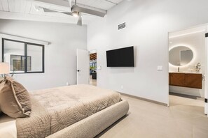 A modern bedroom with a large bed, a flat-screen TV, and a bathroom with a vanity and mirror. - Stunning South Palm Desert Home with Private Pool, Spa & Shadow Mountain Golf Course Views (Palm Desert)