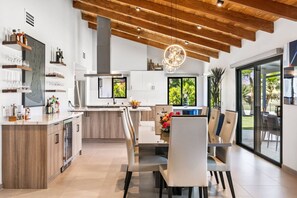 A modern kitchen with a central island, built-in appliances, and a dining area with a large window overlooking a garden. - Stunning South Palm Desert Home with Private Pool, Spa & Shadow Mountain Golf Course Views (Palm Desert)
