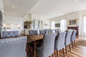 A spacious dining area with a long wooden table and upholstered chairs, a kitchen with white cabinets, and a living area with a fireplace and a television.
