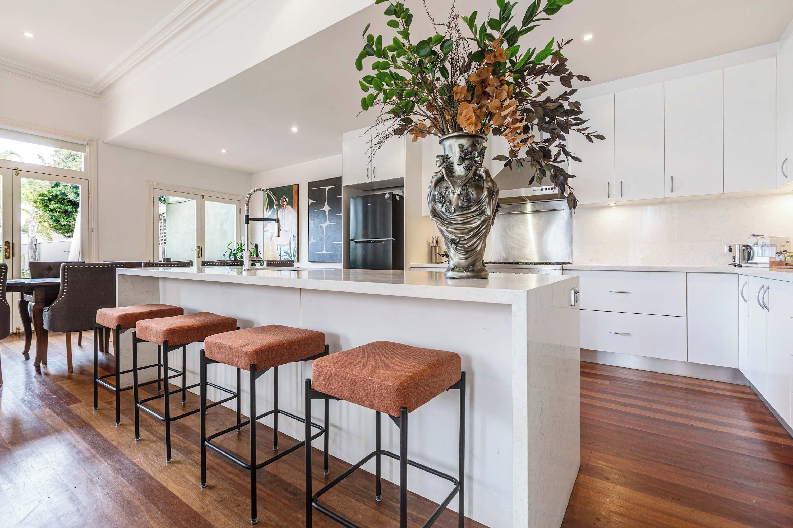 Une cuisine moderne avec des meubles blancs, un îlot central doté de tabourets et un vase décoratif.