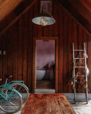 Un interno rustico in legno con un tavolo di legno, una bicicletta, una lampada a sospensione e una scala con piante essiccate.