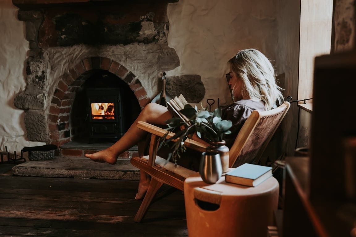 Une personne est assise près d’une cheminée en pierre, un livre et une plante à portée de main, dans une pièce au plancher de bois et à l’ambiance rustique.