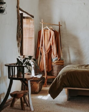 Una camera da letto con un letto, un armadio in legno, un appendiabiti con vestiti, un tavolino con una pianta e un cesto in vimini.