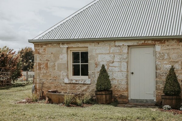 A stone building with a metal roof, a single window, and a door. There are two potted plants in front of the door.