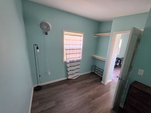 Ein Zimmer mit türkis-blauen Wänden, einem Standventilator, einem Fenster mit gestreifter Vorhang und einem Holzboden. Eine Tür führt in ein weiteres Zimmer.