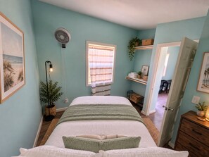 Ein Schlafzimmer mit einem Bett, einem Standventilator, einem Fenster mit Vorhängen, einer Pflanze und einer Tür zu einem anderen Raum.