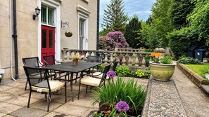 Um pátio externo com mesa e cadeiras, uma porta vermelha e um jardim com diversas plantas e flores.