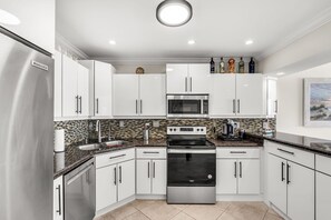 Cozinha moderna com armários brancos, eletrodomésticos de aço inoxidável, painel de azulejos e iluminação embutida no teto.