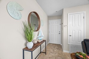 Een hal met een witte deur, een houten console tafel met een potplant, decoratieve wandkunst en een klein model zeilboot.