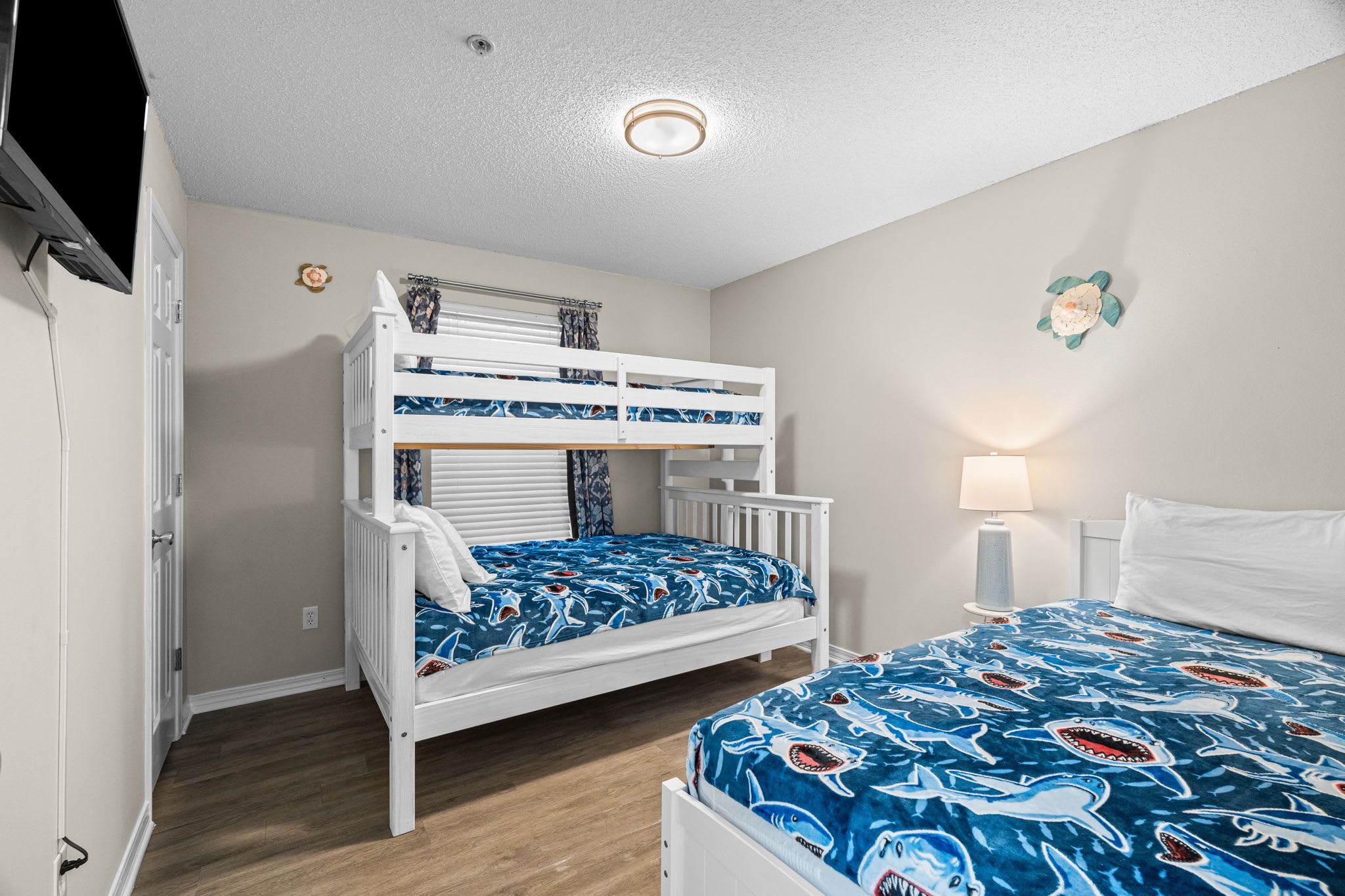 A bunk bed with a blue shark-themed bedspread, a bedside table with a lamp, and a white door.