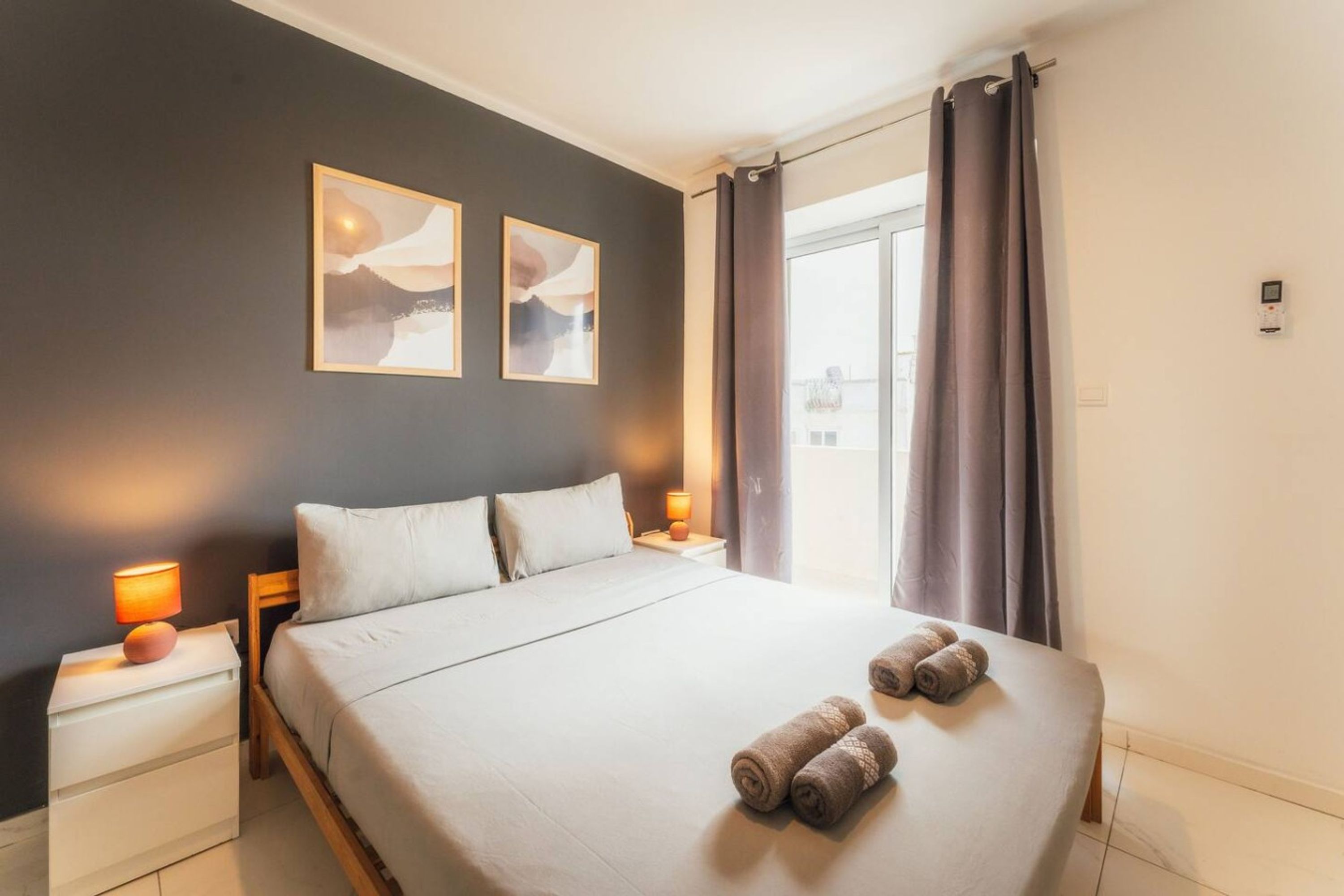 Una habitación de hotel con una cama, dos mesitas de noche, un ventanal con cortinas y dos obras de arte colgadas en la pared.
