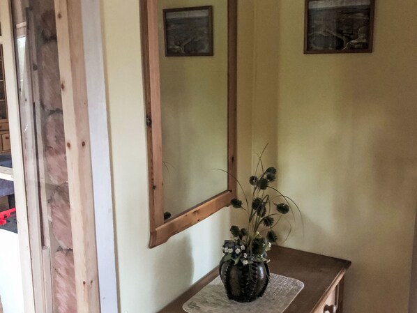 A room with a wooden-framed mirror on the wall, a small table with a vase of flowers, and a picture frame above the mirror.