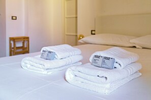 Cama bem arrumada com toalhas brancas dobradas e duas caixas de artigos de higiene pessoal.