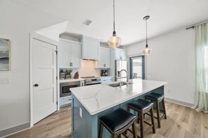 A modern kitchen with a central island, bar stools, pendant lighting, and a view to the living area. - Downtime Panama Palms | Pool, Beach, Pet-Friendly (Panama City Beach)