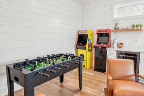 Ein Raum mit einem Kicker, zwei Spielautomaten, einem braunen Ledersessel und einer Küchenzeile inklusive Mikrowelle.