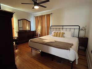 Une chambre d’hôtel avec un lit, un plancher en bois, un ventilateur de plafond, une table de chevet avec une lampe et une commode.