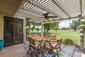 Eine überdachte Terrasse mit Tisch und Stühlen, Blick auf einen Golfplatz und ein Gebäude mit strukturierter Fassade.