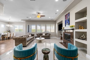 A spacious living room with a high ceiling, a ceiling fan, a fireplace, and a large window. - Lago Vista Retreat | Pool & Game Room | Half Court Basketball | 4BR (Indio)