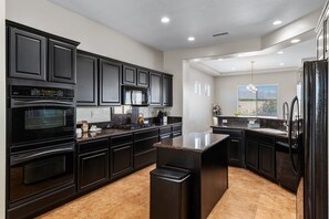 A modern kitchen with dark cabinetry, a central island, and a view of the outdoors through a window. - Lago Vista Retreat | Pool & Game Room | Half Court Basketball | 4BR (Indio)