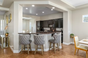 A modern kitchen with a central island, bar stools, dark cabinetry, and a dining area with a white table and chairs. - Lago Vista Retreat | Pool & Game Room | Half Court Basketball | 4BR (Indio)