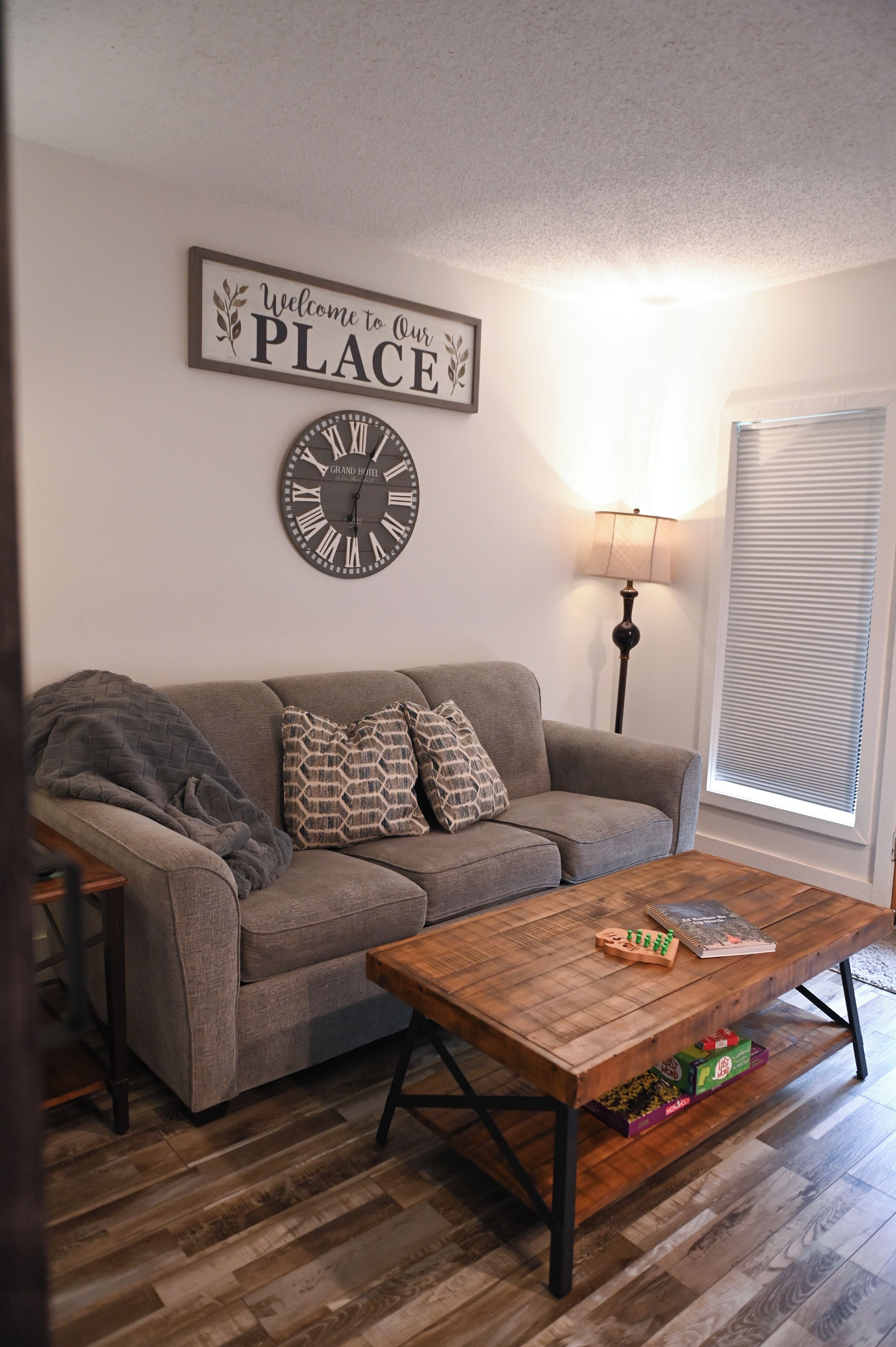 A living room with a grey sofa, patterned cushions, a wooden coffee table with books, a wall clock, a lamp, and a window with blinds.