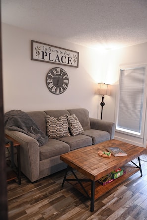 Ein Wohnzimmer mit grauem Sofa, gemusterten Kissen, einem Holztisch mit Büchern, einer Wanduhr, einer Lampe und einem Fenster mit Jalousien.