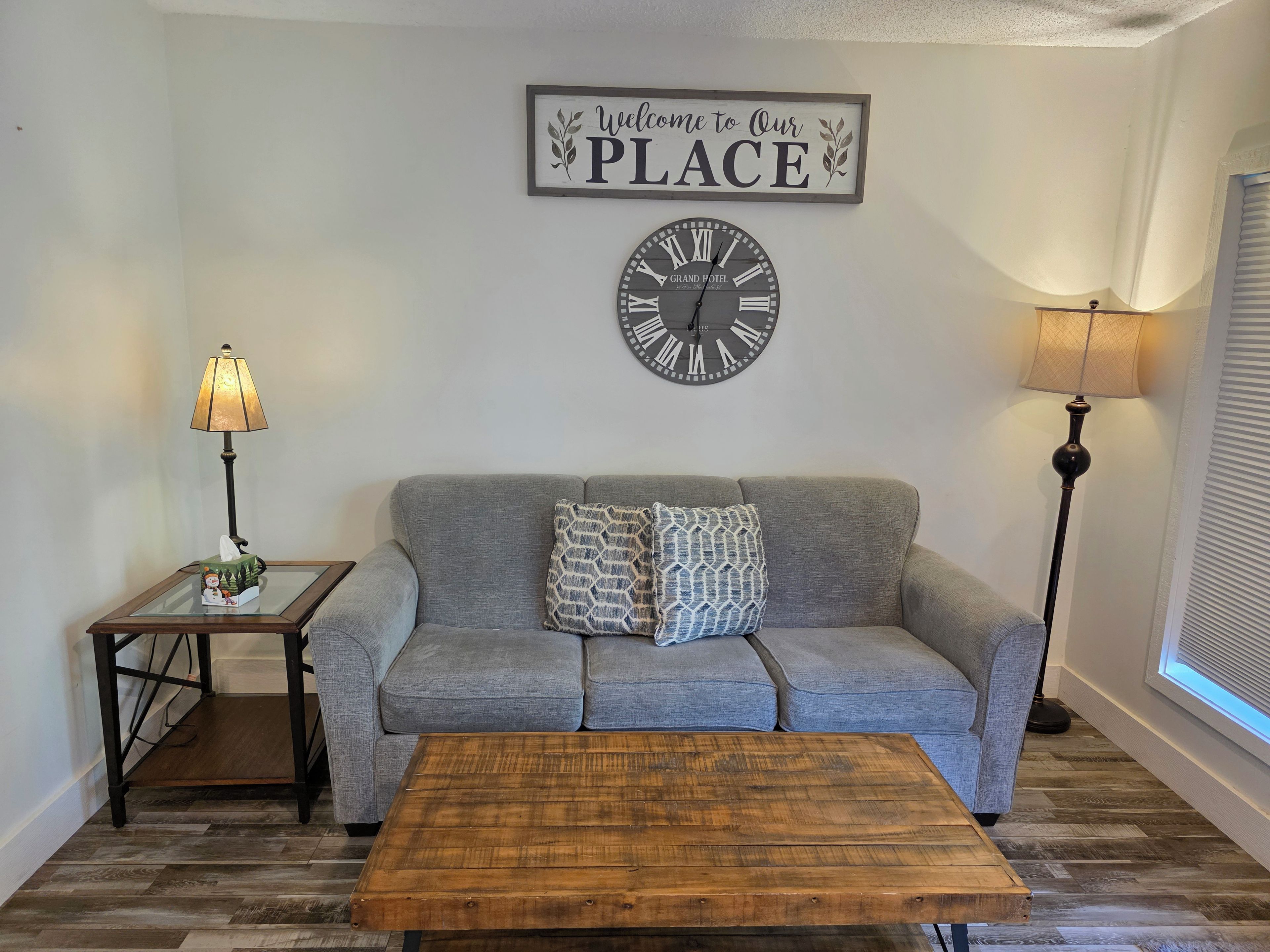 Un salon avec un canapé gris, une table basse en bois, une horloge murale, une lampe et un panneau d’accueil.