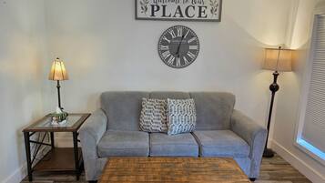 Un salon avec un canapé gris, une table basse en bois, une horloge murale, une lampe et un panneau d’accueil.