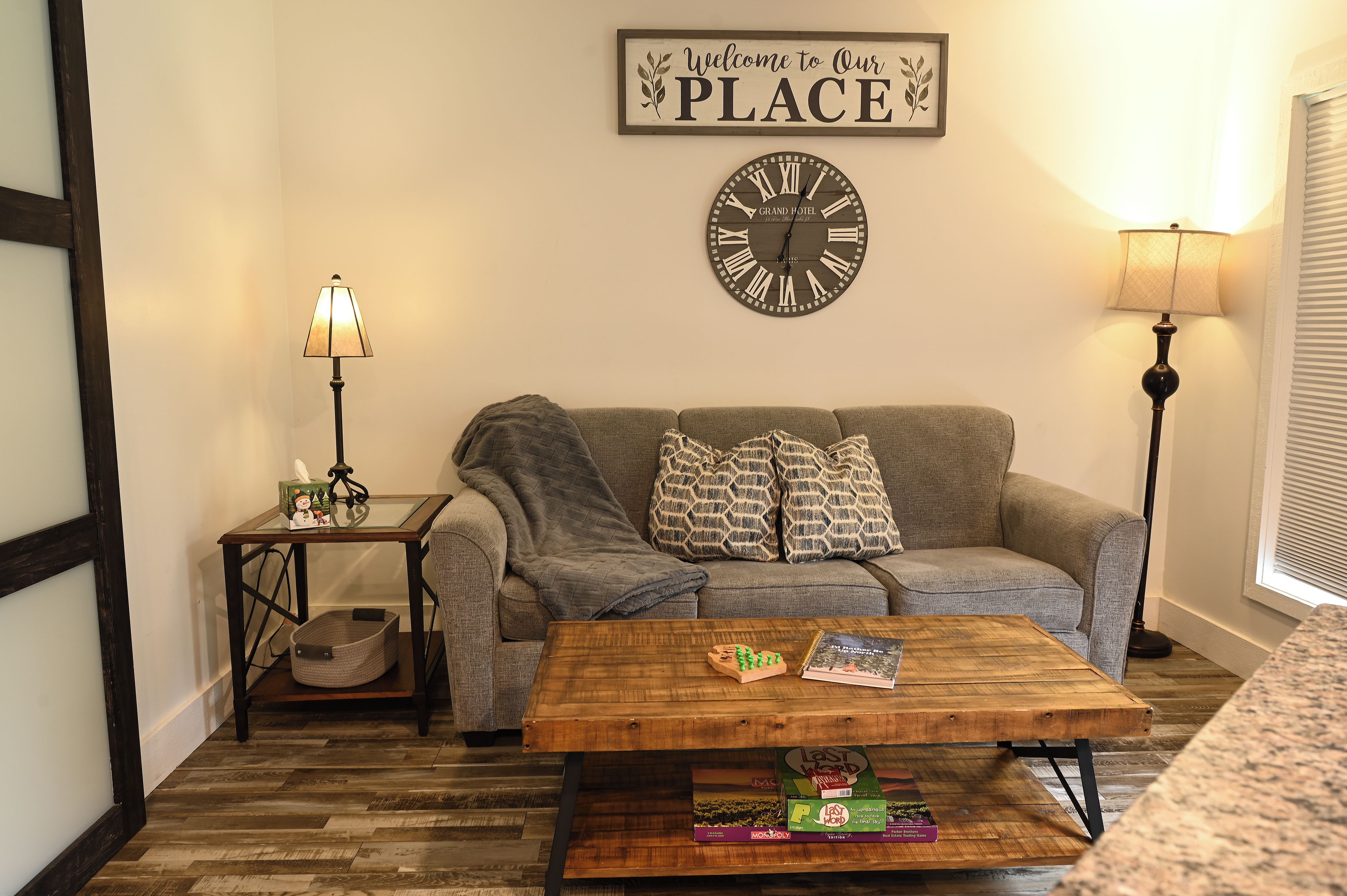 A living room with a couch, a coffee table, a wall clock, and a sign that reads 'Welcome to Our PLACE'.