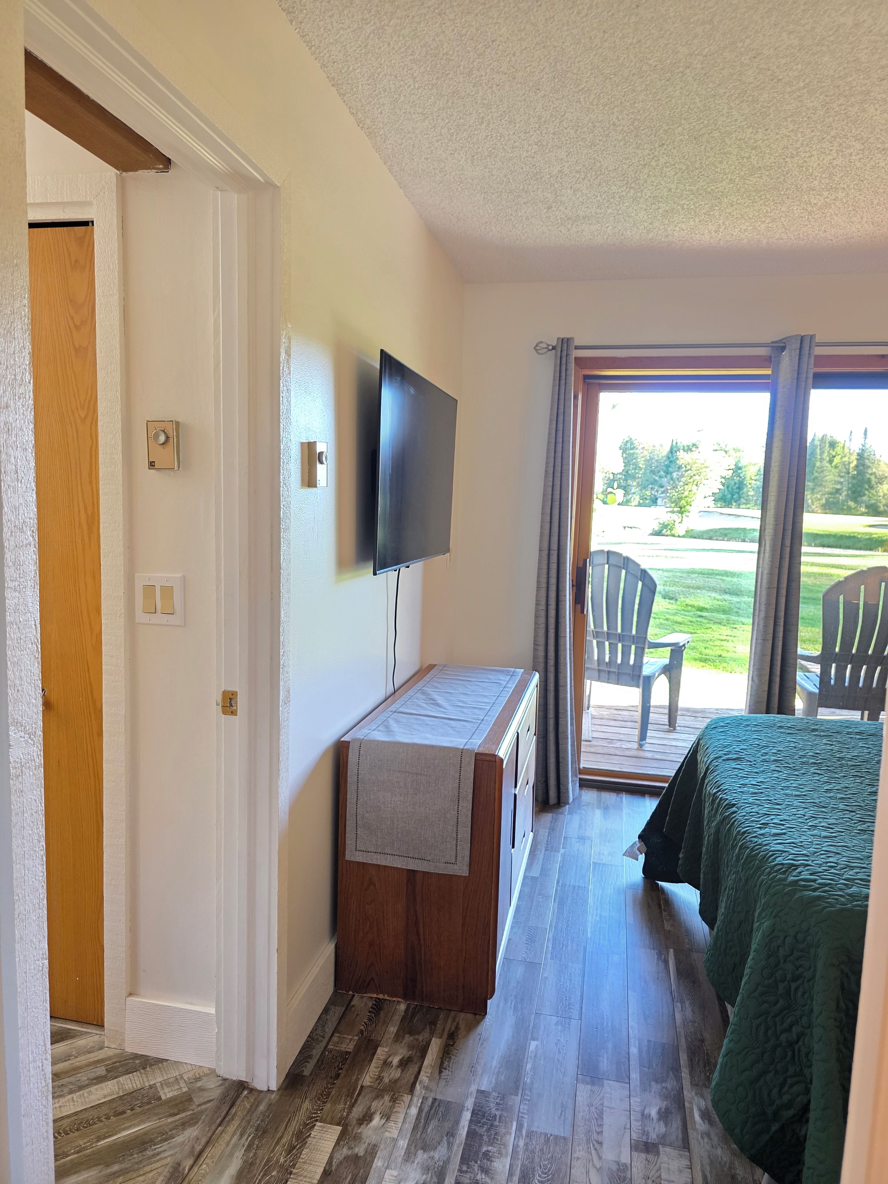 A room with a bed, a TV mounted on the wall, a wooden desk, and a view of the outdoors through a sliding door.