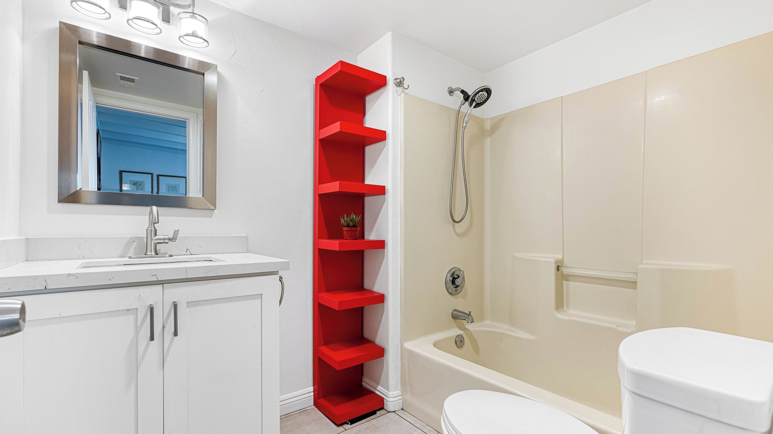 A bathroom with a white sink, a mirror, a red shelf, a shower, and a toilet.