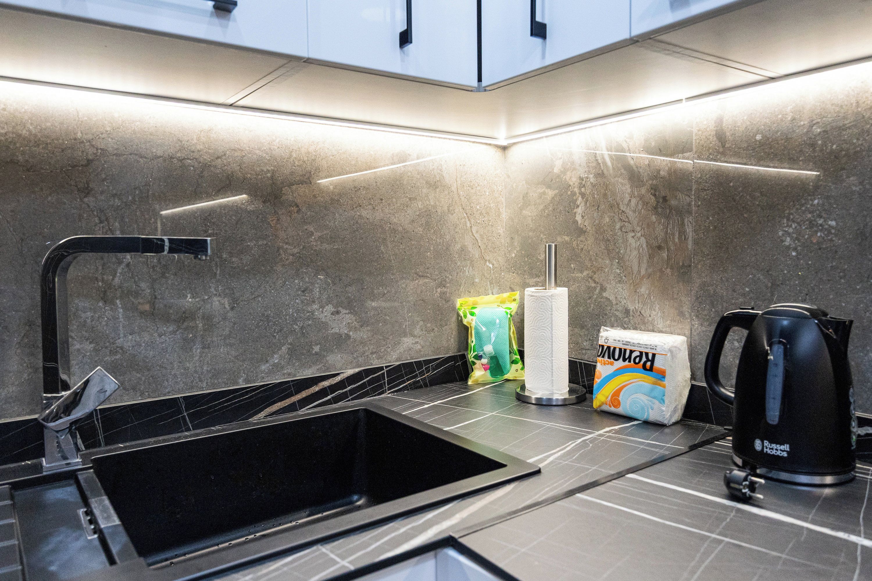 A modern kitchen with a black sink, a kettle, and cleaning supplies.