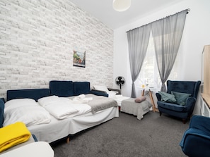 Ein modernes Zimmer mit einer Ziegelwand, einem Bett mit weißer Bettwäsche, einem blauen Sessel, einem kleinen Tisch mit einem Ventilator und einem Fenster mit grauen Vorhängen.