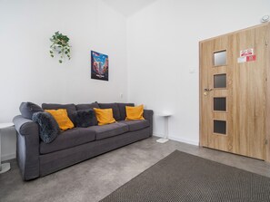 Ein modernes Wohnzimmer mit einem grauen Sofa, einem kleinen Beistelltisch, einer Holztür mit einem Sprechloch und einem gerahmten Stadtbild an der Wand.