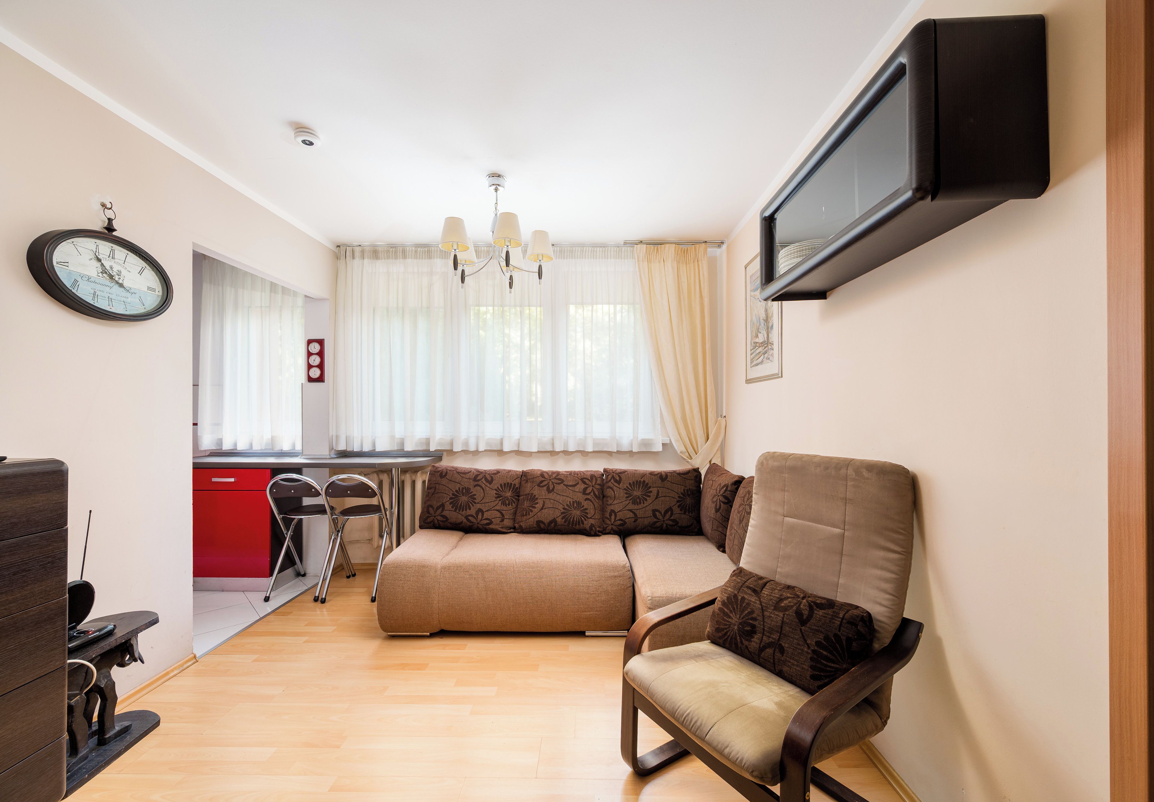 Een woonkamer met een bank, een aan de muur bevestigde televisie, een klok en een raam met gordijnen.