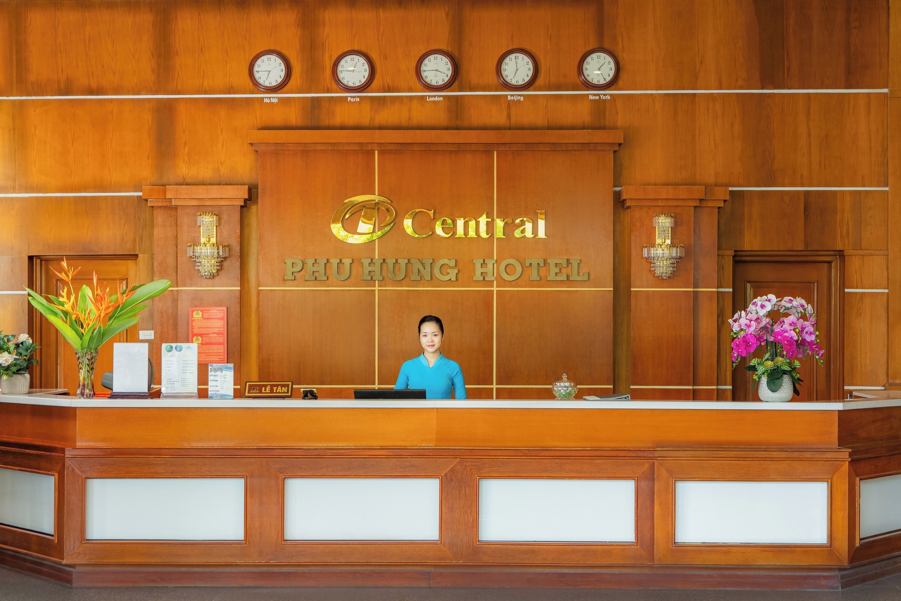 A hotel reception desk with clocks displaying times from various cities, a receptionist, and floral arrangements.