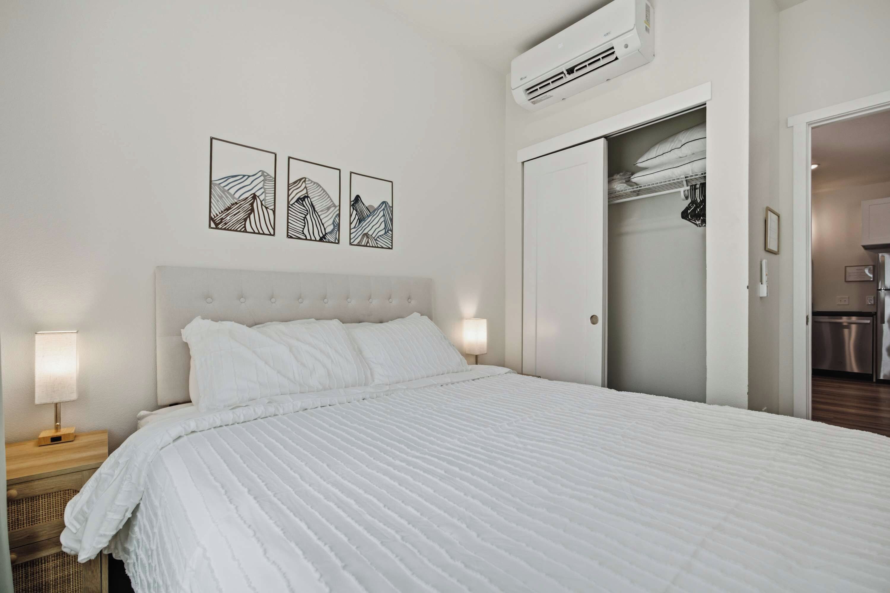 Una cama bien hecha con edredón blanco, una lámpara de noche y un aire acondicionado de pared.