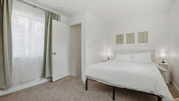 Un dormitorio con cama, mesita de noche, lámpara, puerta y ventana con cortinas.