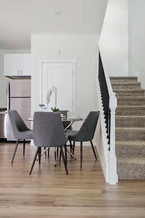 A modern kitchen with white cabinets, a double-door refrigerator, and a dining area with a glass table and two grey chairs. - Sweetheart City Downtown Retreat | Dog Friendly (Loveland)