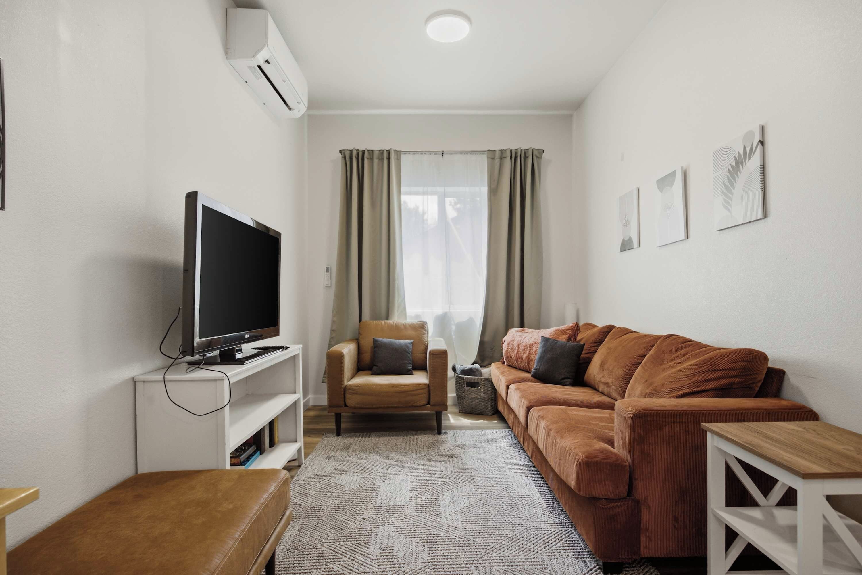 Un espace de vie compact comprenant un téléviseur à écran plat posé sur un meuble, un canapé en cuir marron, une petite table en bois et un tapis à motifs.