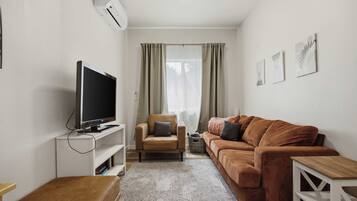 Un espace de vie compact comprenant un téléviseur à écran plat posé sur un meuble, un canapé en cuir marron, une petite table en bois et un tapis à motifs.