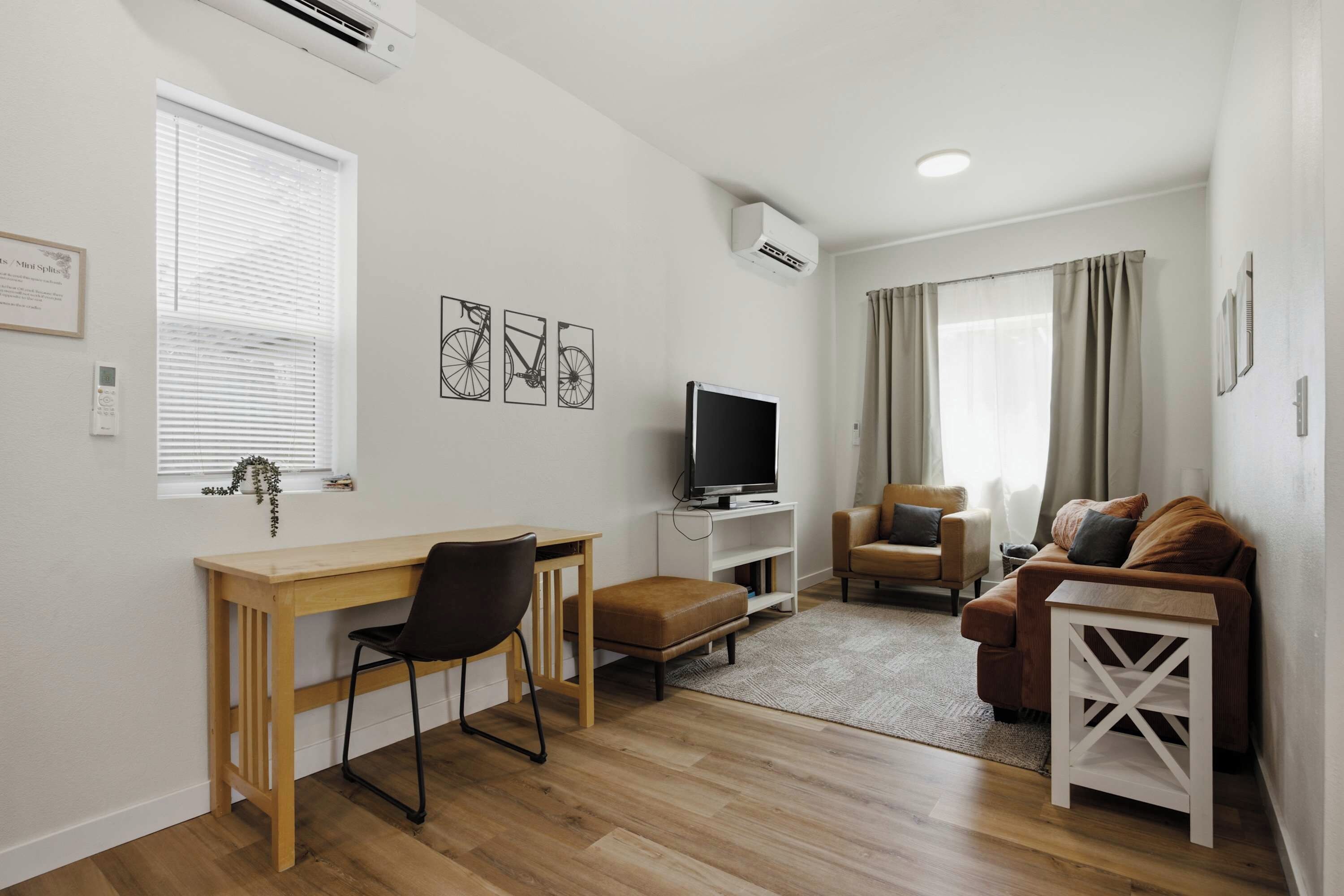 A compact living space with a wooden desk, a television, a sofa, and a window with blinds.