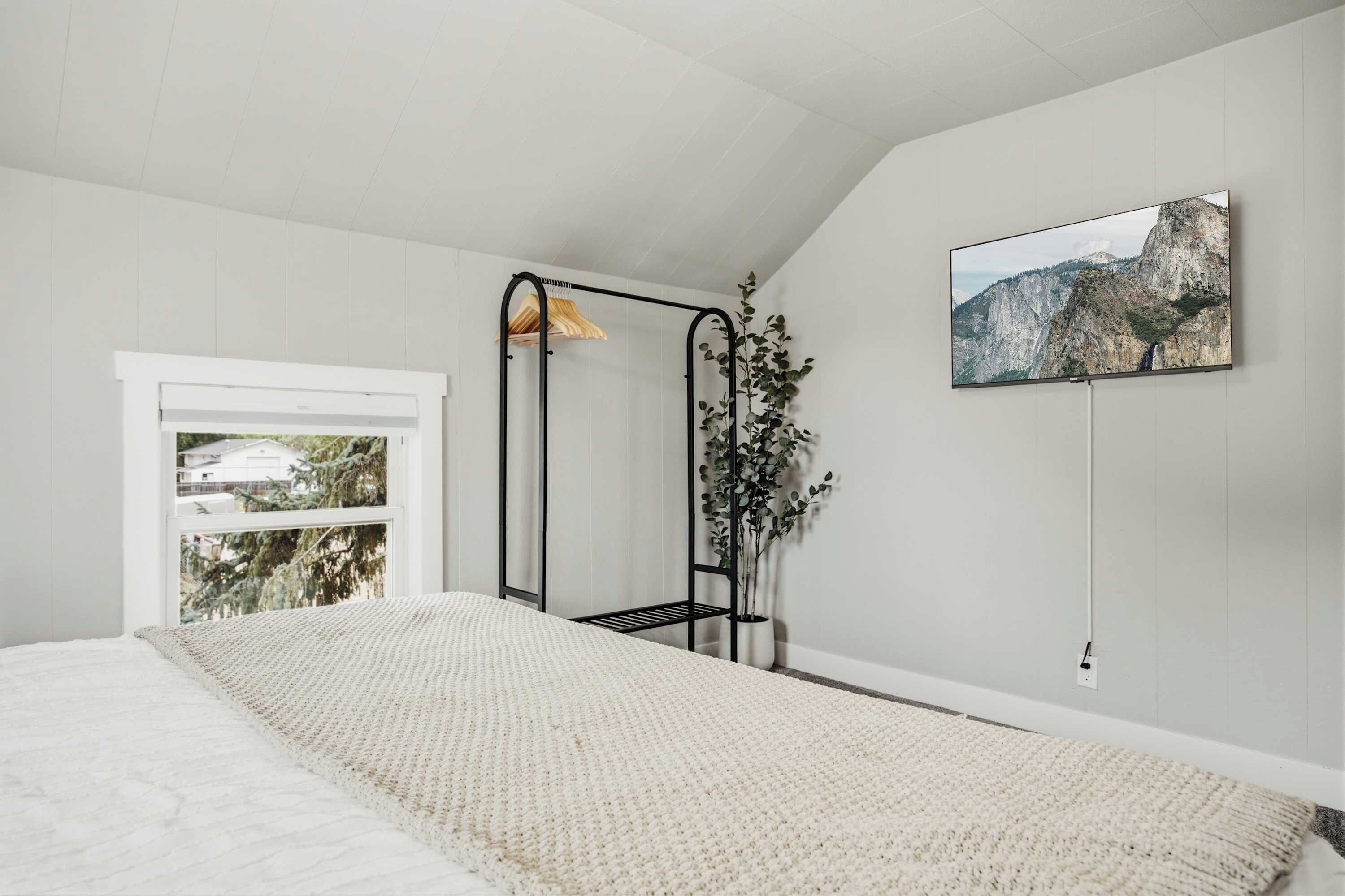 A bedroom with a bed, a window, a plant, and a painting on the wall.
