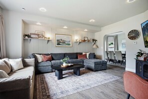 A modern living room with a sectional sofa, a coffee table, and a rug. There is a dining area with a table and chairs visible in the background. - The Evergreen Home | King Beds + Fire Pit (Loveland)