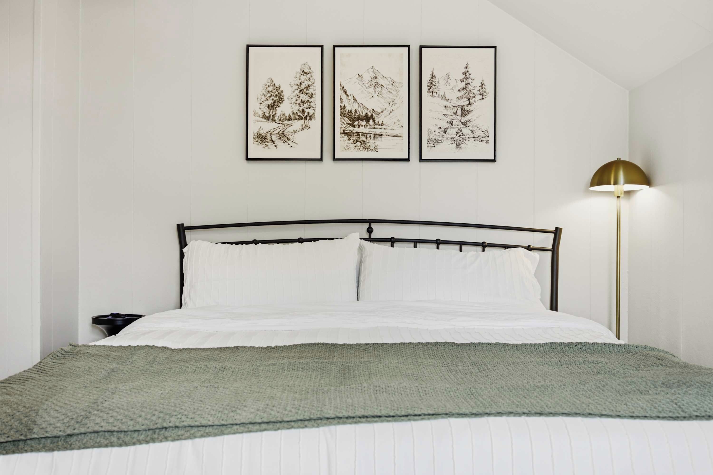A bed with a metal frame, white bedding, and a green blanket. There are three framed artworks above the bed.