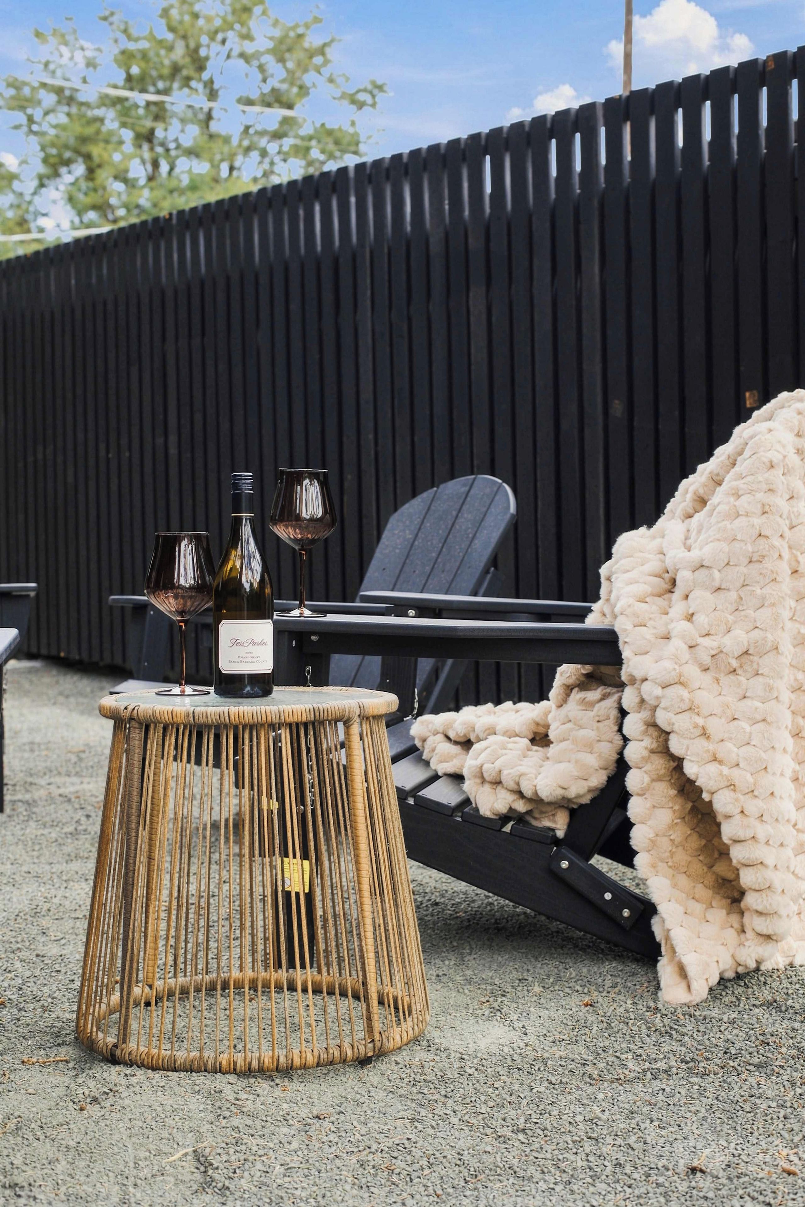 A wicker table with wine bottles and glasses, a black lounge chair, and a beige blanket on a gravel surface.