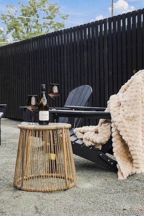 Une table en osier avec des bouteilles de vin et des verres, un fauteuil noir et une couverture beige posés sur un sol recouvert de gravier.