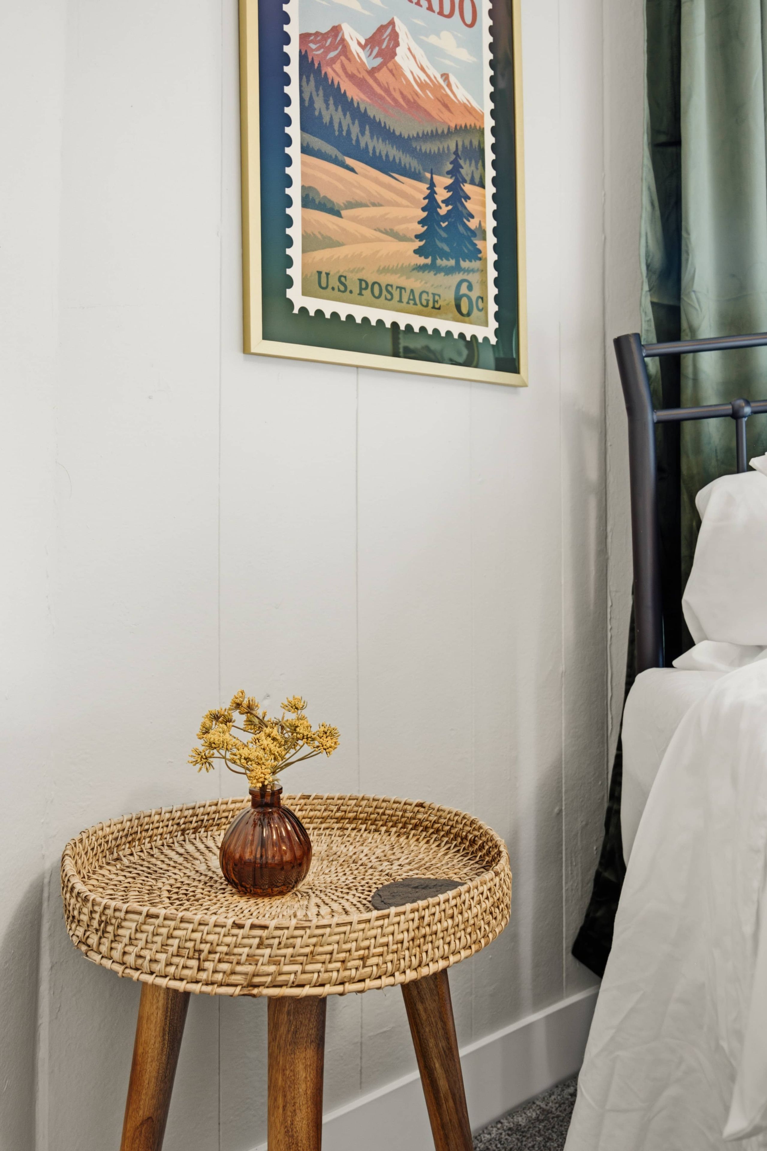 Una habitación con un sello postal enmarcado en la pared, una mesa redonda tejida con un jarrón de flores secas y una cama con cubrecama blanca.